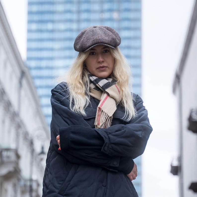 Puede incluir: Una mujer con una gorra de conductor marr&oacute;n, una chaqueta negra y una bufanda de cuadros crema y roja. Est&aacute; de pie frente a un edificio alto.
