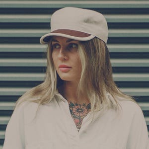 May include: A cream-colored cap with a brown brim. The cap is worn by a person with blonde hair and a white shirt. The background is a striped pattern of dark and light blue.