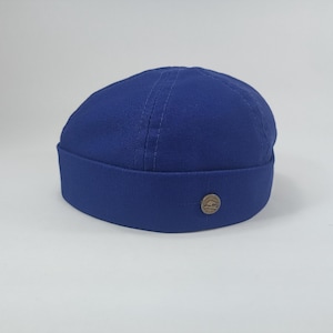 May include: A royal blue docker hat with a rounded crown and a short, folded brim. The hat has a small, circular, bronze-coloured emblem on the brim. The hat is made of a durable fabric and is suitable for casual wear.