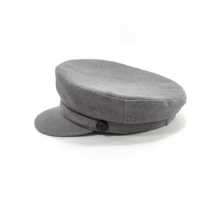 May include: A gray, flat-brimmed cap with a rounded crown and a small, curved visor. The cap features a dark band and a black button detail. The hat is made of a textured fabric and is set against a white background.