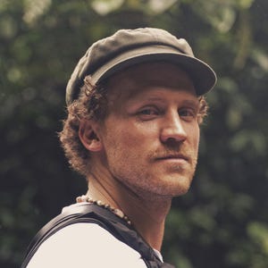 Op de afbeelding: Een man met een groene pet en een wit shirt. Hij heeft krullend bruin haar en een snor. Een kralenketting hangt om zijn nek. De achtergrond is een vage groene bladermassa.