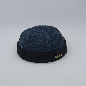 May include: A blue and black wool beanie with a herringbone pattern and a black ribbed cuff. The beanie has a small brown leather label with the word "Hendricks" on it.