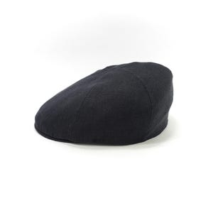 May include: A black, flat cap made of a textured fabric. The hat is a classic style, with a rounded crown and a small, stiff brim. The cap is set against a white background.