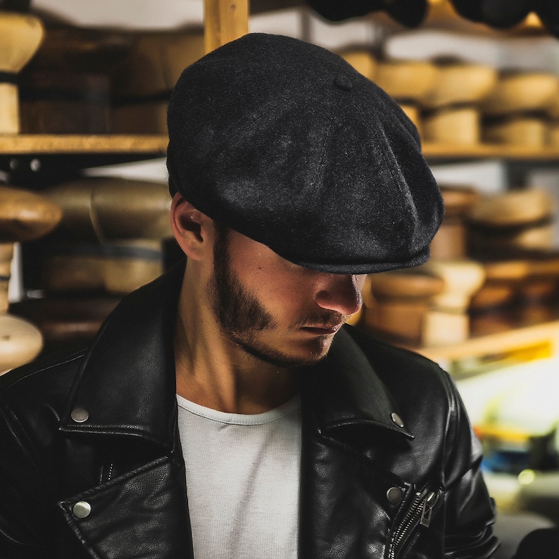 May include: Close-up view of a dark charcoal gray wool newsboy cap. The cap features a classic eight-panel design with a slightly curved brim.  The fabric appears to be soft and textured. The cap is shown against a blurred background of wooden shelves filled with various hats.