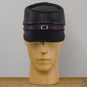 May include: A black leather cap with a short brim and a decorative strap with a silver buckle. The cap features red stitching along the seams and on the strap. The hat is displayed on a wooden mannequin head.