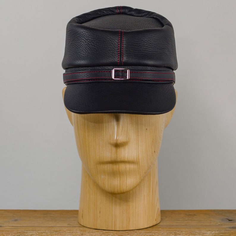 May include: A black leather cap with a short brim and a decorative strap with a silver buckle. The cap features red stitching along the seams and on the strap. The hat is displayed on a wooden mannequin head.