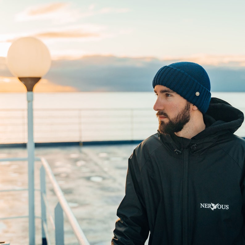 May include: A man wearing a blue beanie and a black jacket with the word "NERVOUS" embroidered on the left side. He is standing on a deck overlooking a body of water.