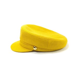 May include: A bright yellow, military-style hat with a small brim and a gold-colored button. The hat has a structured design and a band detail around the crown. The hat is made of a textured fabric.