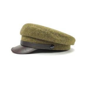 Op de afbeelding: Olijfgroene wollen pet met een bruin leren klep en band. De pet heeft een ronde kroon en een kleine, zwarte knoop aan de zijkant. Een modieus accessoire.