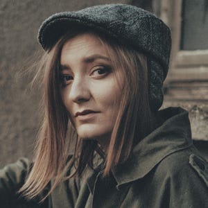 May include: A person wearing a dark grey tweed newsboy cap and a green jacket. The person has long, light brown hair and is looking towards the camera. The background is a weathered, textured wall.