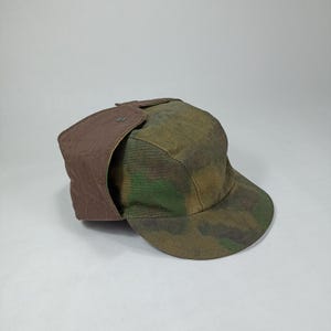 Pode incluir: Um chapéu de camuflagem marrom e verde com uma aba de orelha marrom. O chapéu tem uma aba curva e um botão no topo.