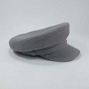 May include: A gray, structured hat with a rounded crown, a short, stiff brim, and a decorative band. The hat features a button detail on the side. The hat is made of a textured fabric.