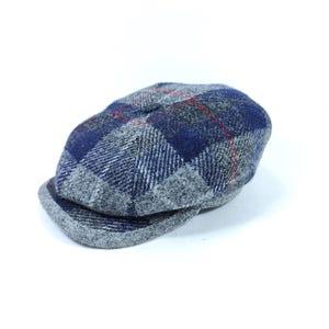 May include: A tweed newsboy cap in a grey, blue, and red plaid pattern. The cap has a rounded crown and a small peak. The fabric has a textured finish, and the plaid design is consistent across the cap. The background is plain white.