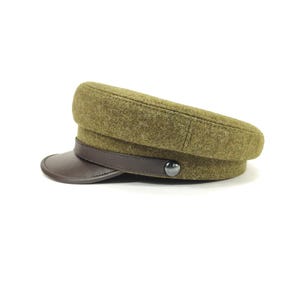 Op de afbeelding: Olijfgroene pet in militaire stijl met een bruin leren vizier en band. De pet heeft een ronde kroon en een knoopdetail aan de zijkant. De hoed is gemaakt van een textuurstof.