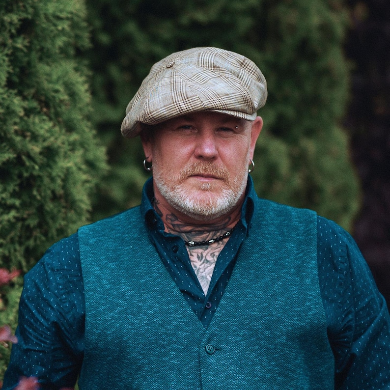 Pu&ograve; includere: Una persona con un berretto a quadri beige, un gilet verde acqua e una camicia blu scuro abbottonata. La persona ha barba, tatuaggi e una collana. Lo sfondo &egrave; un fogliame verde sfocato.