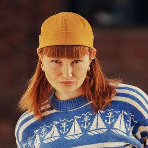 May include: Mustard yellow beanie hat. The hat is worn by a person with red hair and freckles. The person is wearing a blue and white striped sweater with a nautical design.