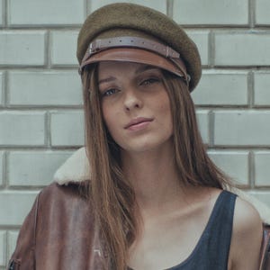 Op de afbeelding: Een vrouw draagt een bruine leren pilotenjack met een crèmekleurige schapenvachtkraag en een donkergroene baret-stijl hoed met een bruin leren klep en riem. De hoed heeft een gespsluiting.