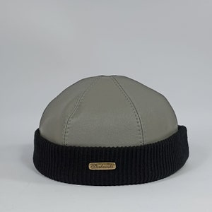 May include: A grey leather beanie with a black knit brim and a gold label that reads "Sterkowski".