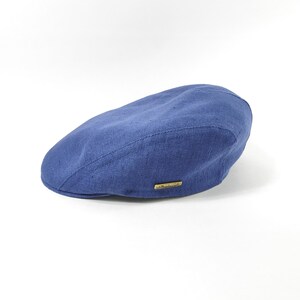 May include: A blue flat cap made of a textured fabric. The cap has a curved brim and a small, rectangular gold-colored emblem on the side. The hat is a classic style, suitable for various occasions.