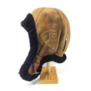 May include: A brown and tan aviator hat with black fur trim, reminiscent of vintage flying gear. The hat has a leather strap and is displayed on a wooden stand. The hat's design suggests warmth and protection.
