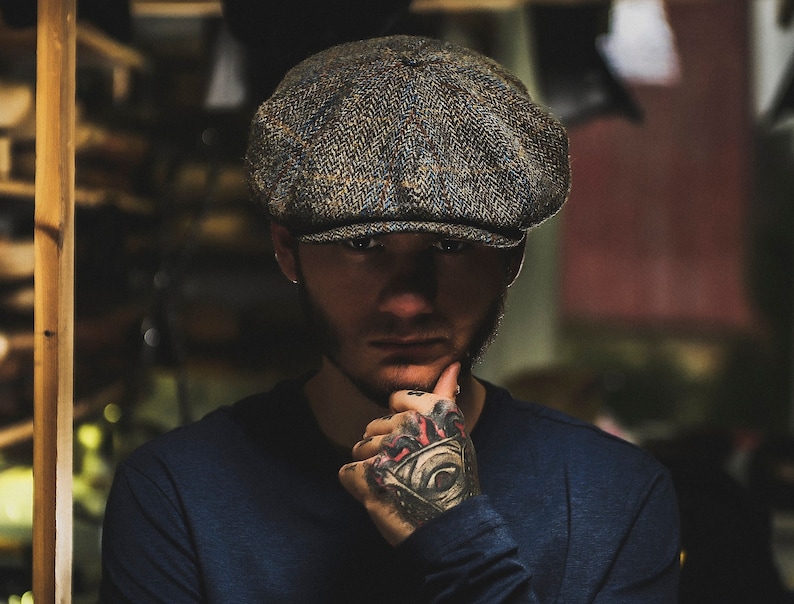 Puede incluir: Un hombre con barba lleva una gorra de tweed de estilo newsboy. Est&aacute; mirando a la c&aacute;mara y tiene la mano en la barbilla. La gorra es marr&oacute;n, azul y gris con un patr&oacute;n de espiga.