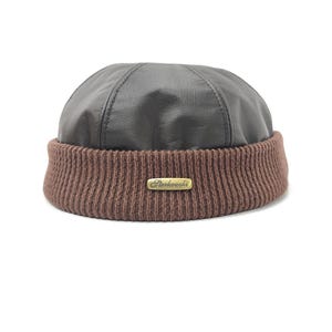 Op de afbeelding: Een bruine leren beanie-muts met een geribbelde gebreide rand. De bovenkant van de hoed is gemaakt van donkerbruin leer en de rand is een lichtere bruine brei. Een klein goudkleurig rechthoekig label met het woord "Sterkowski" is op de rand genaaid.