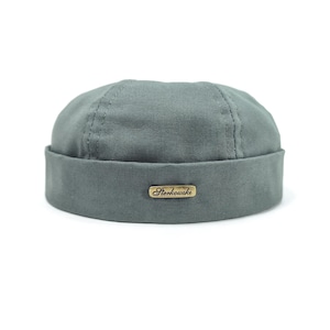 Puede incluir: Un gorro tipo docker de color verde salvia hecho de tela de lona resistente. El gorro presenta una corona redondeada con costuras visibles y un ala ancha doblada. Una pequeña placa metálica rectangular color latón está fijada al frente con el texto Sterkowski.