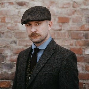 May include: A man wearing a brown tweed newsboy cap, a blue button-down shirt with a white collar, a patterned vest, and a dark gray tweed suit jacket.