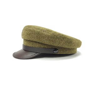 Op de afbeelding: Een olijfgroene pet in militaire stijl met een donkerbruin leren vizier en band. De pet heeft een afgeronde kroon en een knoopdetail aan de zijkant. De hoed staat tegen een witte achtergrond.