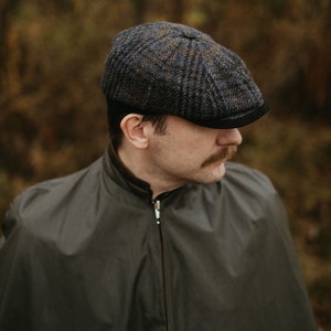 May include: A man wearing a dark green jacket with a zip and a tweed newsboy cap with a black brim. The cap has a blue, brown, and grey pattern.