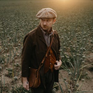 K&ouml;nnte beinhalten: Ein Mann tr&auml;gt eine braune Jacke, eine braune Weste, eine braune Ledertasche und eine braune und beige gemusterte Flatcap. Er steht auf einem Feld mit gr&uuml;nen Pflanzen.