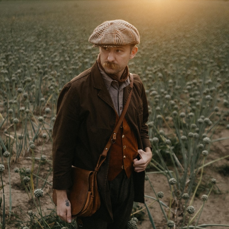 K&ouml;nnte beinhalten: Ein Mann tr&auml;gt eine braune Jacke, eine braune Weste, eine braune Ledertasche und eine braune und beige gemusterte Flatcap. Er steht auf einem Feld mit gr&uuml;nen Pflanzen.