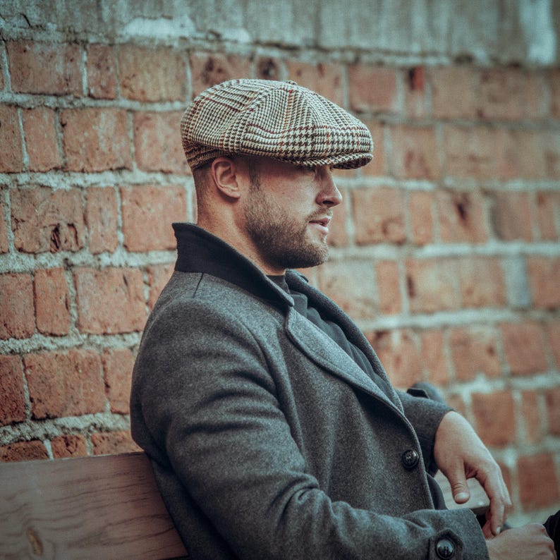K&ouml;nnte beinhalten: Ein Mann mit einer braunen und schwarzen karierten Newsboy-M&uuml;tze sitzt auf einer Holzbank vor einer Backsteinmauer. Er tr&auml;gt einen dunkelgrauen Mantel und ein schwarzes Hemd.