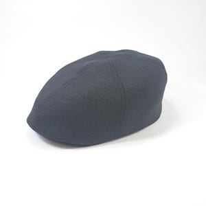 Peut inclure: Casquette plate gris foncé, avec une couronne arrondie et une petite visière rigide. Le chapeau est fait d'un tissu texturé et posé sur un fond blanc. Ce style classique convient à diverses occasions.