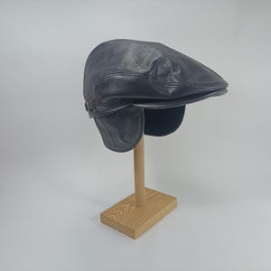 以下が含まれることがあります： 木製のスタンドに展示された、濃いグレーの革製のフラットキャップ。 キャップは、湾曲したつばと側面のボタンのディテールを備えたクラシックなデザインです。 耳当ては、暖かさを増すために着用できます。