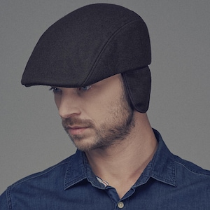 May include: A dark gray newsboy cap with ear flaps. The cap is made of a soft, textured material and has a classic, rounded shape. The ear flaps extend down to cover the ears. The cap is worn by a person.