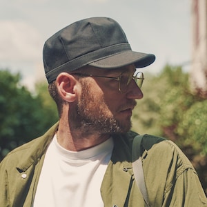 May include: A dark charcoal gray cap with a short, structured brim and a rounded crown. The cap features a clean, minimalist design with a stitched band detail above the brim, crafted from a solid, matte fabric material.