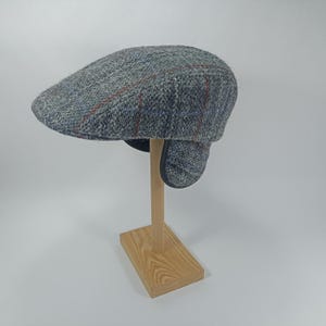 May include: A grey tweed flat cap with ear flaps, displaying a plaid pattern with red and blue detailing. The hat is presented on a wooden stand, highlighting its traditional design and textured fabric. A classic accessory for colder months.