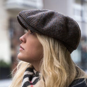 Puede incluir: Una mujer con una gorra de estilo newsboy de color marr&oacute;n y azul con una visera marr&oacute;n. La gorra est&aacute; hecha de tejido de tweed y tiene una corona ligeramente redondeada.