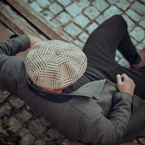 K&ouml;nnte beinhalten: Eine Person tr&auml;gt eine graue Tweedjacke und eine schwarze Hose mit einer braunen und wei&szlig;en karierten Newsboy-M&uuml;tze.