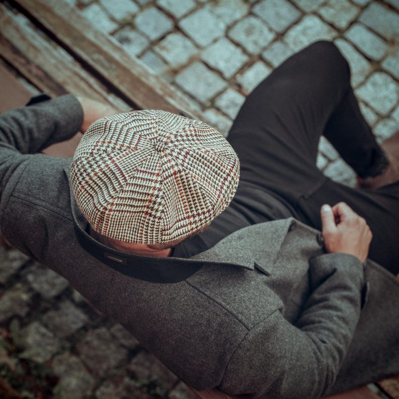 K&ouml;nnte beinhalten: Eine Person tr&auml;gt eine graue Tweedjacke und eine schwarze Hose mit einer braunen und wei&szlig;en karierten Newsboy-M&uuml;tze.
