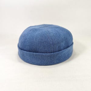 May include: A denim blue cap with a rolled brim. The hat is made of a textured fabric and sits on a white surface. This casual hat is a stylish accessory.