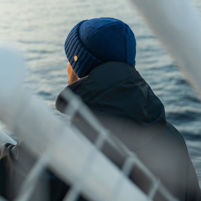 May include: A person wearing a dark blue beanie and a dark blue jacket, standing on a boat with a white railing. The person is looking out at the water.