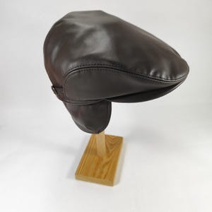 Può includere: Un cappello newsboy in pelle marrone scuro con paraorecchie. Il cappello è esposto su un supporto di legno su sfondo bianco. Il cappello ha una corona arrotondata e una piccola visiera rigida. I paraorecchie sono attaccati ai lati e possono essere ripiegati.