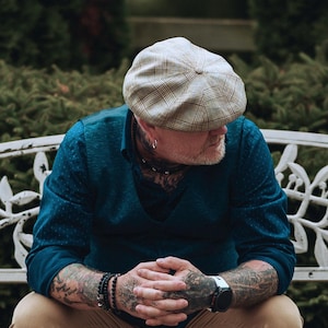 May include: A person wearing a plaid newsboy cap, a dark teal button-down shirt, and a black watch. The person has tattoos on their arms and neck. They are sitting on a white bench with a decorative design, with green foliage in the background.