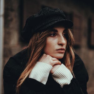 May include: A woman with long brown hair is wearing a black newsboy cap and a black jacket. She is looking off to the side and has her hands resting on her chin.