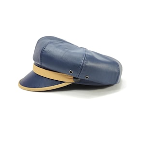 Può includere: Un berretto in stile marinaio blu navy realizzato in materiale liscio con corona strutturata e pannelli cuciti. Il berretto presenta una fascia beige a contrasto sul davanti, una visiera con bordo beige e due piccoli occhielli in metallo scuro.