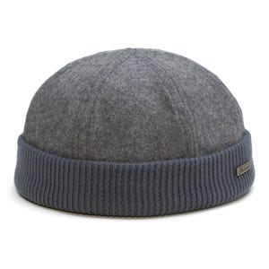 May include: A grey felted wool docker cap featuring a structured six-panel crown and a wide, ribbed dark blue knit cuff. A small, rectangular metallic tag with engraved text is attached to the side of the cuff, providing a subtle detail.