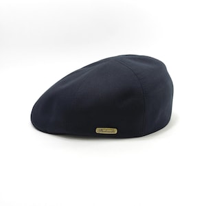May include: A navy blue flat cap with a curved brim. The cap has a smooth texture and a small, rectangular gold-colored label on the side. The cap is a classic style, suitable for various occasions.