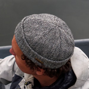 May include: A grey and black herringbone patterned cap. The cap is made of a woven fabric and has a short brim. A small, rectangular metal accent is on the side of the cap.